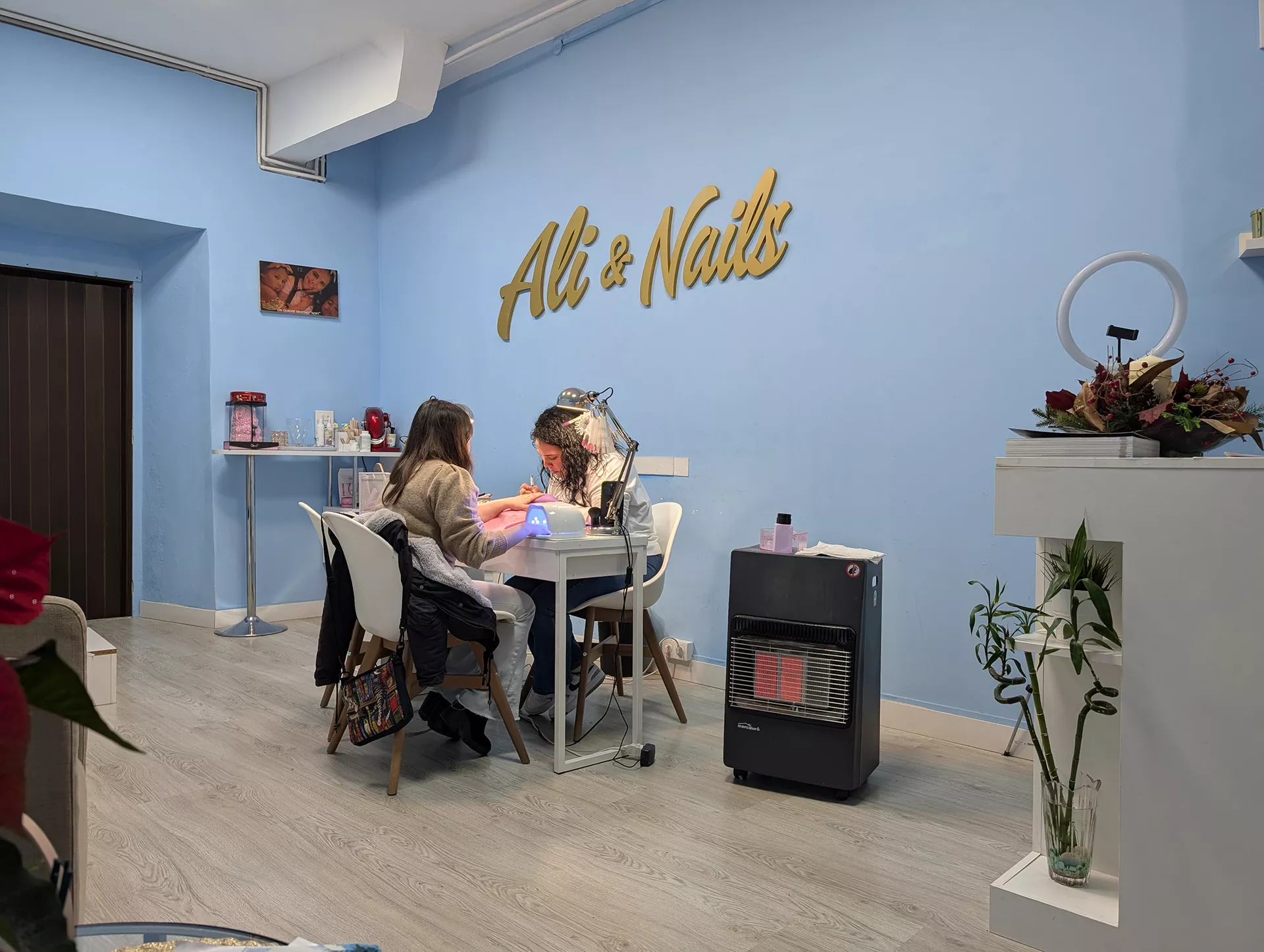 Mujer haciendo trabajo de manicura a otra mujer, cartel de Ali Nails en la pared y decoración